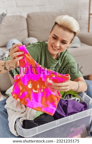 happy emotion, joyful woman sorting second-hand clothes and holding colorful top, trendy hairstyle, tattoo, blurred background, sustainable living and mindful consumerism concept