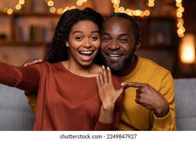 Happy emotional beautiful black couple got engaged on Valentines evening, cheerful man and woman sitting on couch at festive home interior, taking selfie together, showing engagement ring and smiling - Powered by Shutterstock