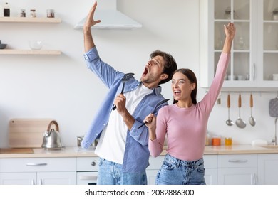 Happy Emotional Funny Millennial European Lady And Guy Have Fun Together, Singing Song At Imaginary Microphone Preparing Food In Kitchen Interior. Active Positive Couple Enjoying Cooking At Free Time