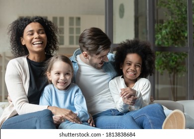 Happy Emotional Mixed Race Family Relaxing Together On Comfortable Couch Indoors. Laughing Multiracial Couple Enjoying Watching Television Funny Films With Joyful Small Biracial Children At Home.