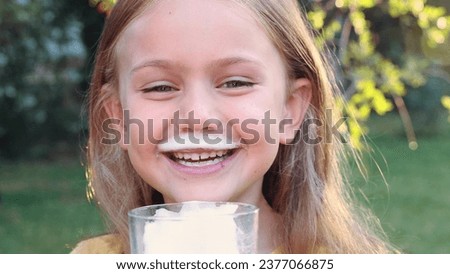 Happy emotional smiling adorable child girl hold glass of milk and drink on nature outside sunny summer morning. Kid open mouth with mustache of milk on lips. Calcium for good children health