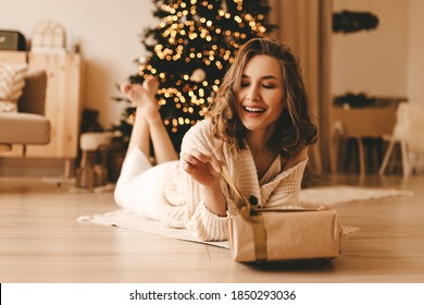 Happy emotional surprised young woman opens a holiday gift sitting on the floor by the Christmas tree in the cozy living room of the house, the concept of happiness