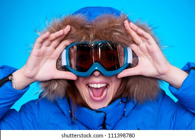 Happy Emotional Woman In Ski Glasses And Winter Coat Over Blue Background