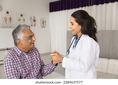 Happy empathetic young doctor touching hand of elderly Indian patient with geriatric diseases, holding arm with support, calming senior man, giving medical care, consultation, smiling, laughing - Powered by Shutterstock