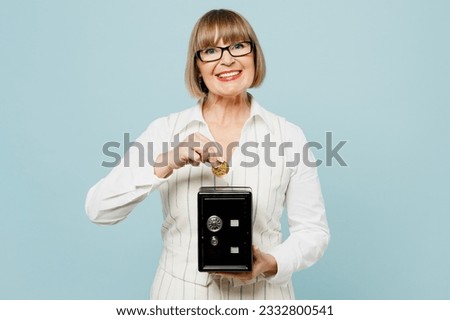 Happy employee business woman 50s wear white classic suit glasses formal clothes hold put coin into metal bank safe for money accumulation isolated on plain blue background. Achievement career concept