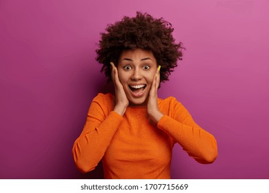 Happy energetic woman grabs face, hears wonderful news, realises she won huge money, stands impressed and speechless, dressed in orange jumper, feels lucky and thrilled, got incredible offer - Shutterstock ID 1707715669