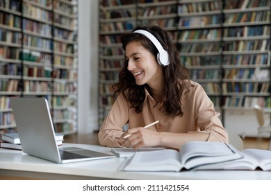 Happy engaged gen Z student girl watching webinar, attending online class, learning conference, talking to teacher on video call, laughing, writing, studying in library with headphones, laptop, books - Shutterstock ID 2111421554