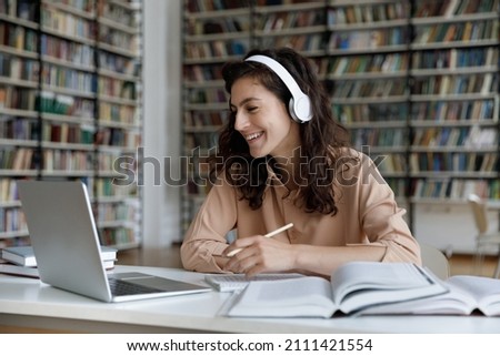 Happy engaged gen Z student girl watching webinar, attending online class, learning conference, talking to teacher on video call, laughing, writing, studying in library with headphones, laptop, books