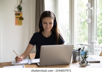 Happy Engaged Young Female Student In Wireless Headphones Watching Learning Webinar, Virtual Class, Online Lesson At Home, Talking To Teacher On Video Call On Laptop, Writing Notes