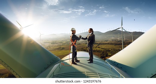 Happy engineer and businessman shake hands after good work. They standing a top of windmill. Around wind generators and beautiful sunset landscape - Shutterstock ID 1243197676