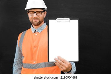 Happy Engineer Pointing At Blank Paper Sheet, Clipboard. Man Builder In Safety Vest And Hard Hat Advertising Empty White Tablet Standing Over Black Background, Mockup. All For Construction. Copy Space