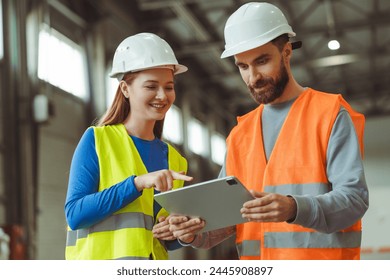 Happy engineers, factory foreman and female worker wearing protective white helmets, using digital tablet, planning startup, standing in warehouse. Cooperation, teamwork concept - Powered by Shutterstock