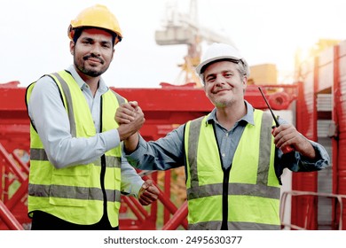 Happy engineers with safety vest and helmet have handshake at building site. Senior elderly foreman shakes hands with young worker during works together at construction site. teamwork and unity. - Powered by Shutterstock