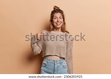 Happy enthusiastic young woman with glad expression smiles broadly points at herself dressed in net jumper and jeans promots herself says choose me isolated over brown background. Hey look at me