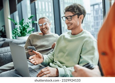 happy entrepreneur in eyeglasses pointing at laptop while working on startup project near business partner with smartphone smiling on blurred background in modern coworking space - Powered by Shutterstock