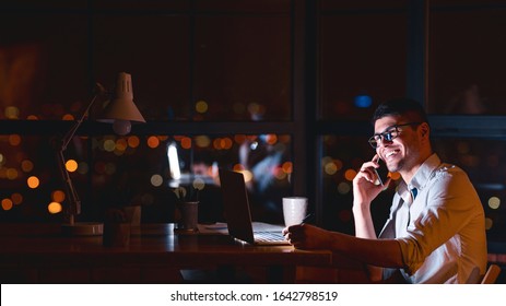 Happy Entrepreneur Guy Talking On Cellphone Sitting At Laptop In Modern Office At Night. Business People Lifestyle. Panorama, Free Space