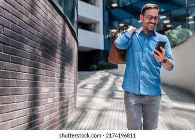 Happy entrepreneur messaging on smart phone. Young professional wearing formals is carrying laptop bag. He is walking on footpath against building during sunny day. - Powered by Shutterstock