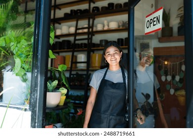 Happy entrepreneur woman wearing an apron working in her plant shop, opening the door and welcoming a customer - Powered by Shutterstock