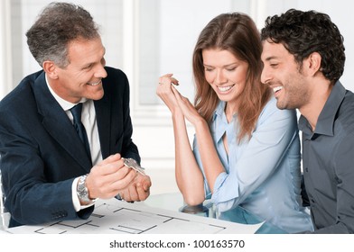 Happy Estate Agent Showing New Home Keys To A Young Couple After A Discussion On House Plans.