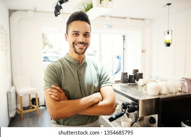 Happy ethnic bartender with crossed arms looking at camera while standing near espresso machine at counter in coffee shop