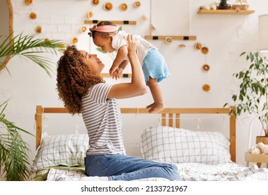 Happy Ethnic  Curly Mom Playing With Laughing Little Baby Daughter In Bed, Playful Little Girl  Flying In Air In Mother's Arms