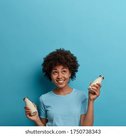 Happy Ethnic Curly Woman Drinks Lactose Free Beverage, Holds Bottle Of Almond Or Coconut Milk, Looks Upwards, Smiles Positively, Isolated Over Blue Background, Copy Space For Your Information