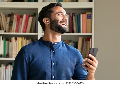 Happy Ethnic Indian Professional Business Man At Work In Home Office Wearing Earbud Using Cell Phone, Smiling Eastern Businessman Having Virtual Mobile Chat Video Call On Smartphone, Watching Game.