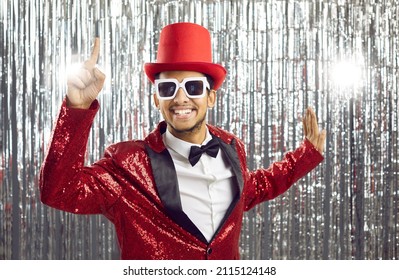 Happy ethnic man in a funny outfit has a great party idea. Excited young guy in a red sequin jacket, tophat and glasses points his index finger up and smiling standing on silver foil fringe background
