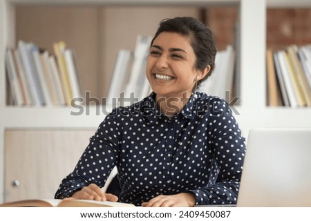 Happy ethnic millennial girl sit at desk in classroom look to side talking with teammate, smiling indian female student take break from studying have fun chatting with group mate or colleague