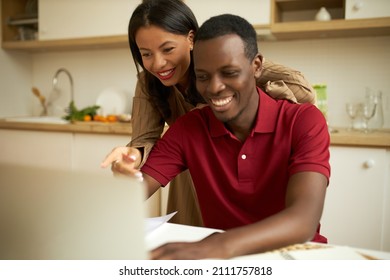 Happy ethnicity couple at kitchen browsing online shops on laptop, smiling, dark-skinned elegant charming woman pointing at screen with cheerful face expression, african man typing with joyful look