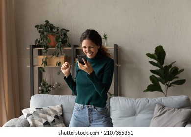 Happy euphoric millennial generation hispanic latin woman looking at mobile phone screen, feeling excited reading message with amazing win news, jumping dancing alone in living room, internet success.