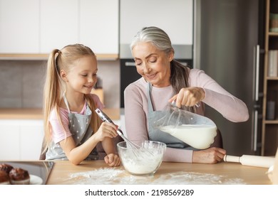 Happy European Elderly Grandmother And Little Granddaughter In Aprons Pour Milk To Dough In Kitchen Interior. Homemade Sweet Food, Cooking Lesson Together For Family At Home, Hobby And Housekeeping