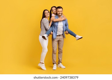 Happy European Family Of Three Having Fun Together Over Yellow Background, Cheerful Young Parents And Their Teen Daughter Laughing And Smiling At Camera, Girl Piggybacking Dad, Copy Space - Powered by Shutterstock