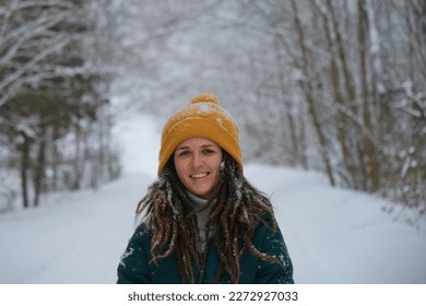 Happy European girl with blue eyes and dreadlocks walks alone in winter park. Close up portrait of young smiling pretty Caucasian woman in yellow knitted hat against background of snowy winter forest.