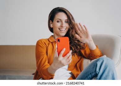 Happy european girl has video call at home. Cheerful young woman is using airpods and smartphone. Wireless earphones using at online conference. Technology using, leisure and relaxation concept.