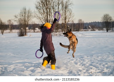 Happy European girl on walk in winter park playing with dog. German Shepherd jumps high in snow in hope of getting blue ring toy. Active games outside.
