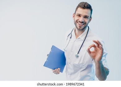 Happy european male doctor hold clipboard and showing Ok gesture. Young bearded man with stethoscope wearing white coat. Isolated on gray background with turquoise light. Studio shoot. Copy space.