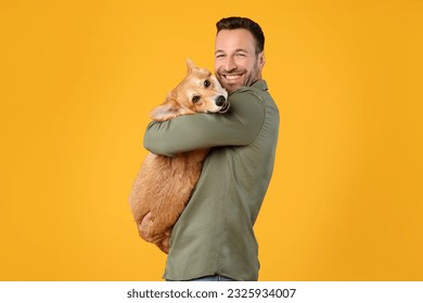 Happy european man holding and comforting cute corgi dog, smiling at camera, posing with pet over yellow studio background