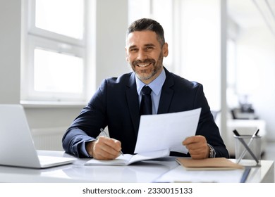 Happy european middle aged businessman working with documents, taking notes to financial report or signing contract, sitting at desk in modern office and smiling at camera