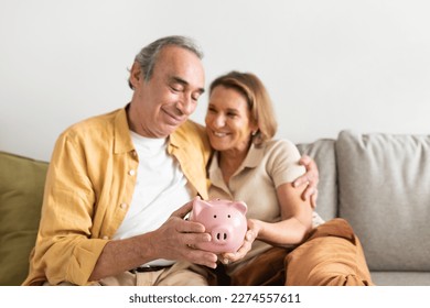 Happy european senior couple holding pink piggybank in hands and embracing, sitting on sofa in living room interior, free space. Retirement savings concept