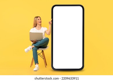 Happy European Young Woman Sit On Chair With Laptop, Pointing Finger At Big Smartphone With Blank Screen, Isolated On Yellow Background, Studio. Online Job, Ad And Offer, Website And App For Study