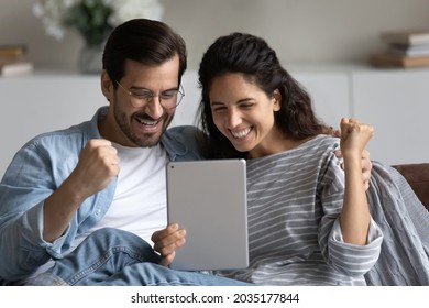 Happy excited 30s couple using tablet at home, looking at screen, reading text, making winner fist yes gesture. Husband and wife celebrating achieve, success, win, getting good surprising news