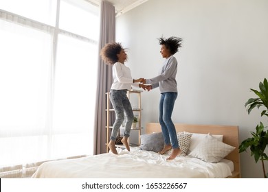 Happy excited african american family mom and cute teen daughter jumping on bed, carefree mixed race mother with teenage girl holding hands play having fun laughing feeling joy fly in air in bedroom
