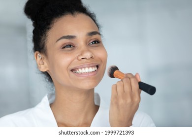 Happy excited African American girl covering face with makeup foundation with toothy smile, enjoying beauty care visage glamour routine, applying powder with big cosmetic brush. Beautician concept