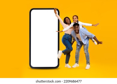 Happy Excited African American Millennial Couple With Little Girl Have Fun Near Big Smartphone With Blank Screen, Isolated On Orange Background, Studio. Ad And Offer, Website And New App For Family