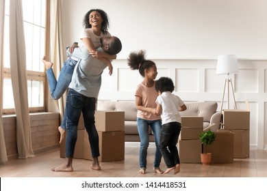 Happy excited african family celebrate moving day relocation together, cheerful black parents embrace and cute kids children jump laugh in living room with boxes, new house purchase and renovation