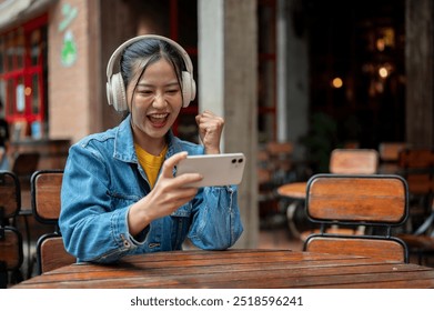 A happy, excited Asian woman in a denim jacket wearing headphones is enjoying playing a mobile game while sitting at an outdoor table of a restaurant.