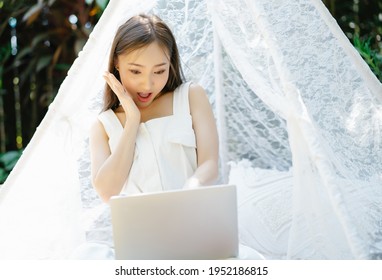 Happy Excited Asian Woman Sit Under Blanket Tent Decorated And Using Laptop Computer With Shocked Expression, Covering Mouth Business Success On Making Big Sale Of Online Store. SME Marketing Online.