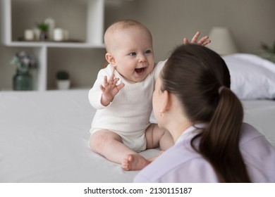 Happy excited baby laughing at mom face, showing positive emotions, waving hands, sitting on bed, playing. New mother talking to charming infant child, cuddling kid, having fun