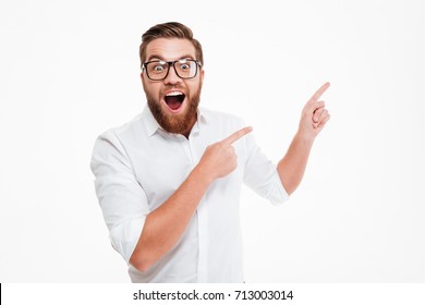 Happy excited bearded man in eyeglasses pointing away at copy space with two fingers isolated over white background - Shutterstock ID 713003014